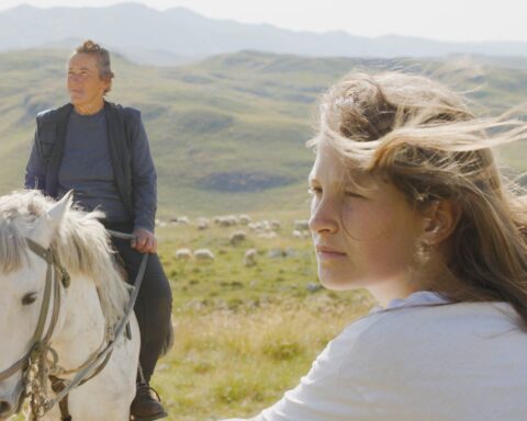 A young woman sits at the foreground of the frame looking off to the left. Her mother is behind her, sitting on a white horse. The rolling pasturelands are in the background.
