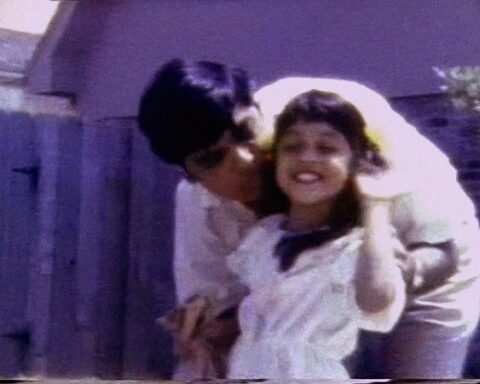 A grainy VHS image of an Indian father hugging and kissing his young mixed race daughter. They are both wearing white shirts. She is waving at the camera.