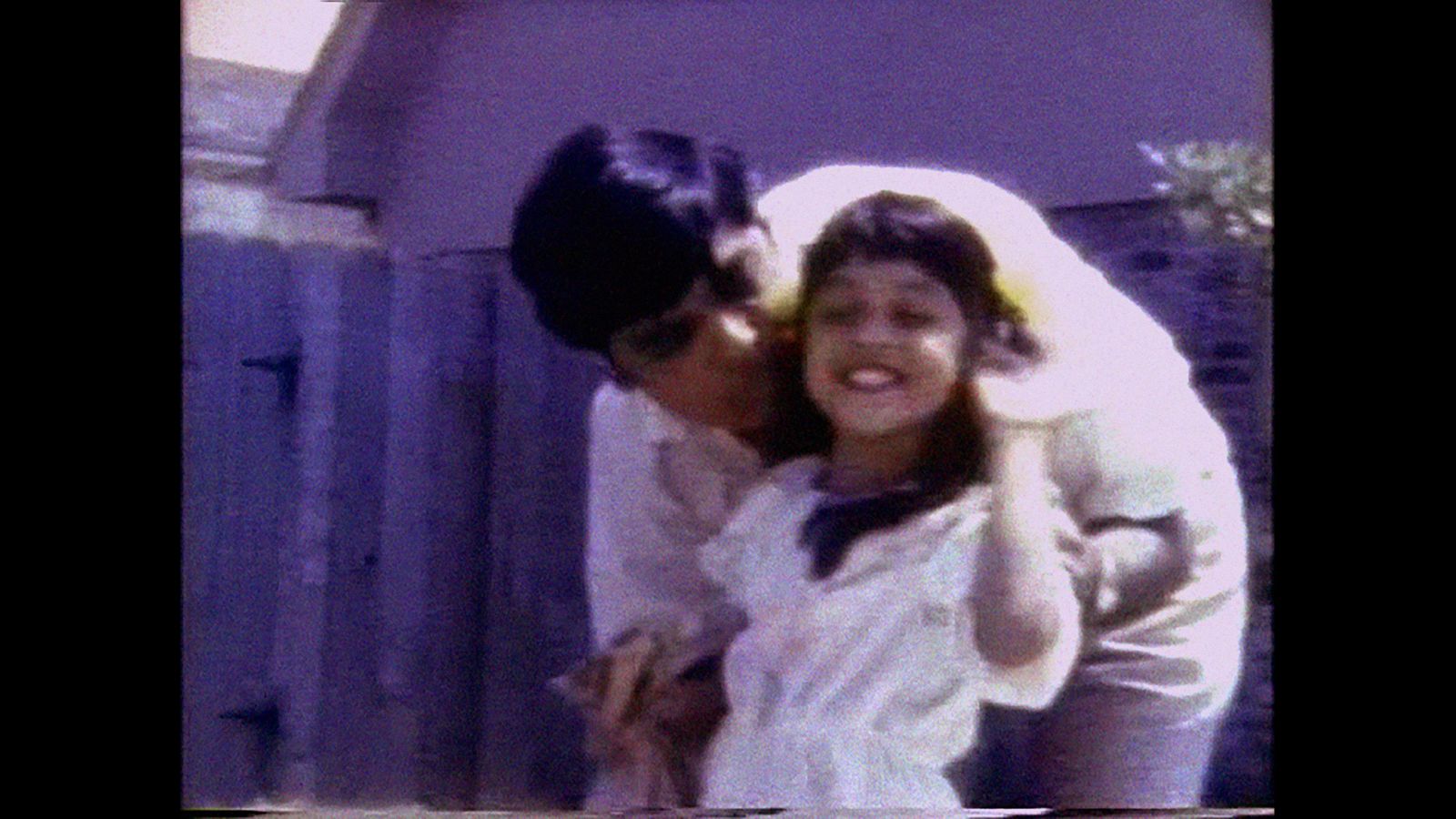 A grainy VHS image of an Indian father hugging and kissing his young mixed race daughter. They are both wearing white shirts. She is waving at the camera.