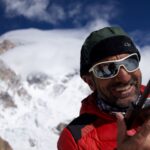 A man in a red winter jacket speaks into a radio. He is standing among the mountain range of K2.