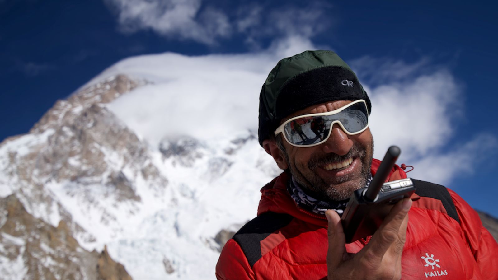 A man in a red winter jacket speaks into a radio. He is standing among the mountain range of K2.