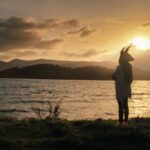 A woman in an Indigenous headdress is pictured in silhouette as she stands at the edge of a river at sunset.