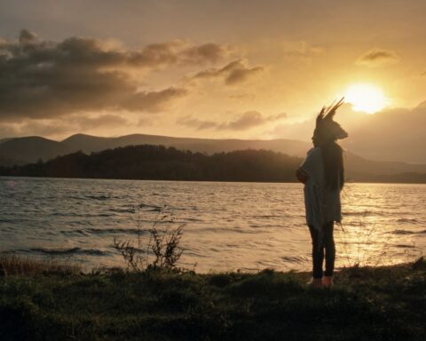 A woman in an Indigenous headdress is pictured in silhouette as she stands at the edge of a river at sunset.