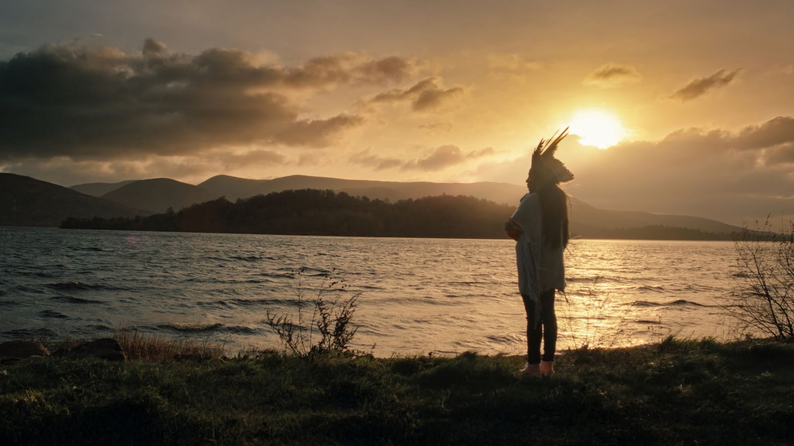 A woman in an Indigenous headdress is pictured in silhouette as she stands at the edge of a river at sunset.