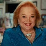 Carol Conners appears seated for an interview. She is smiling and has red hair, bright red lipstick,and is wearing a blue suit.