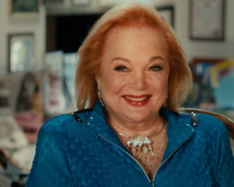 Carol Conners appears seated for an interview. She is smiling and has red hair, bright red lipstick,and is wearing a blue suit.