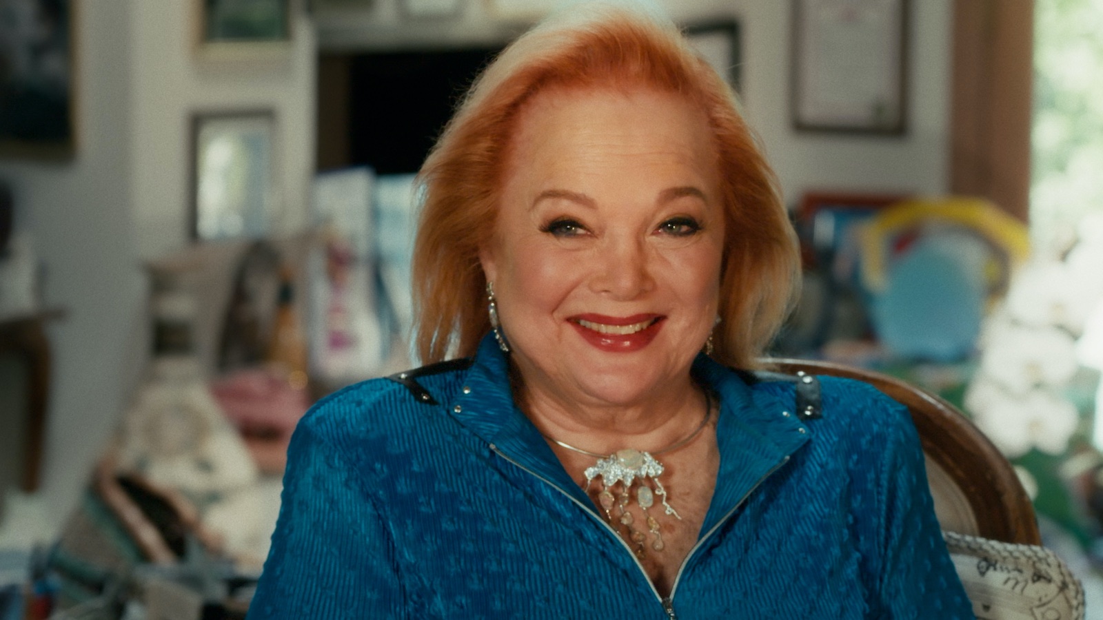 Carol Conners appears seated for an interview. She is smiling and has red hair, bright red lipstick,and is wearing a blue suit.