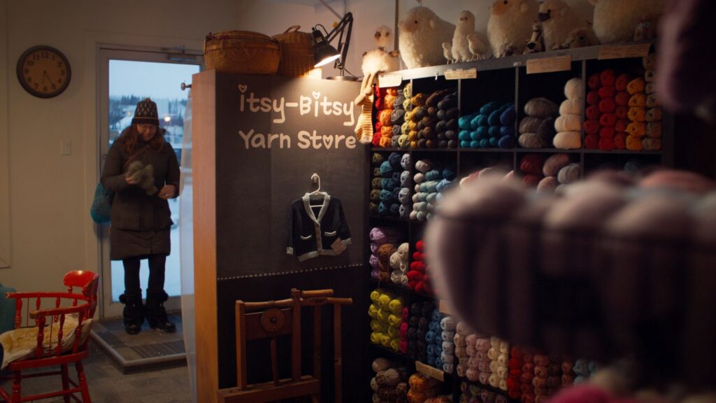 A woman enters a knitting store. There is a chalkboard sign that advertises "The Itsy-Bitsy Yarn Store" and there are shelves of yarn bundles of different.