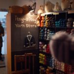 A woman enters a knitting store. There is a chalkboard sign that advertises "The Itsy-Bitsy Yarn Store" and there are shelves of yarn bundles of different.