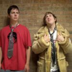 Two male high school students lean against a brick wall. The one on the left is wearing a red t-shirt with a dark striped tie, and the one on the right is wearing a tan trench coat, white shirt, and loose black tie. He is holding the lapels of the coat.