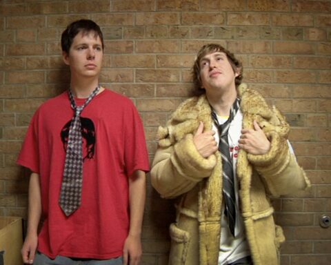 Two male high school students lean against a brick wall. The one on the left is wearing a red t-shirt with a dark striped tie, and the one on the right is wearing a tan trench coat, white shirt, and loose black tie. He is holding the lapels of the coat.
