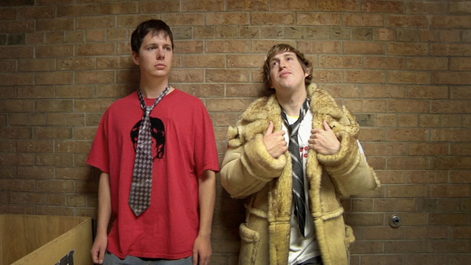 Two male high school students lean against a brick wall. The one on the left is wearing a red t-shirt with a dark striped tie, and the one on the right is wearing a tan trench coat, white shirt, and loose black tie. He is holding the lapels of the coat.