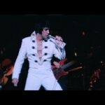 Elvis Presley performs in concert wearing a whte jumpsuit.