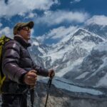 A Sherpa stands looking at Mount Everest. He is wearing a baseball cap and sunglasses and is holding two ski poles.