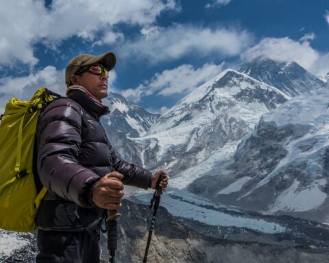 A Sherpa stands looking at Mount Everest. He is wearing a baseball cap and sunglasses and is holding two ski poles.