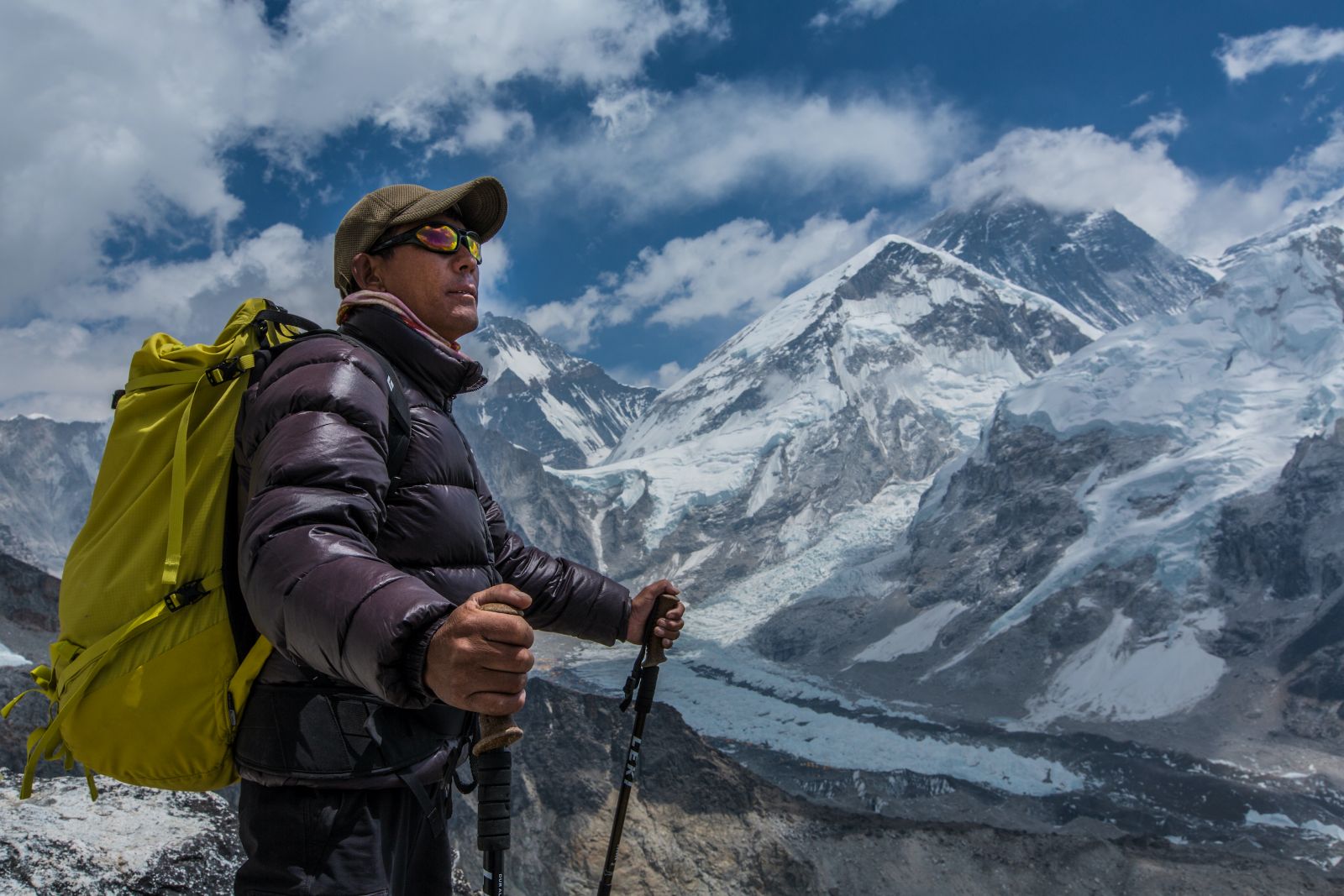 A Sherpa stands looking at Mount Everest. He is wearing a baseball cap and sunglasses and is holding two ski poles.
