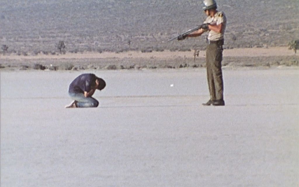 A man kneels on the ground while a police officer stands above him and aims a shotgun at him. They are in barren area.