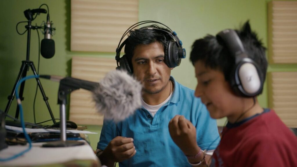 A Peruvian father and his young son sit at a microphone. They are both wearing headphones as the father directs his son in a vocal performance.