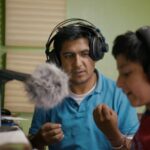 A Peruvian father and his young son sit at a microphone. They are both wearing headphones as the father directs his son in a vocal performance.