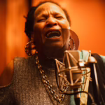 Reggae singer Sister Nancy performs behind a microphone in a recording studo. Her eyes are cosed as she sings wiith emotion. The background of the studio is a golden orange colour.