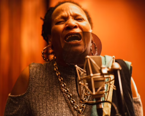 Reggae singer Sister Nancy performs behind a microphone in a recording studo. Her eyes are cosed as she sings wiith emotion. The background of the studio is a golden orange colour.