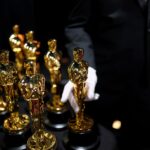 Two rows of Oscar statuettes stand backstage at the 2025 Academy Awards. A pair of hands in white gloves holds one statuette.