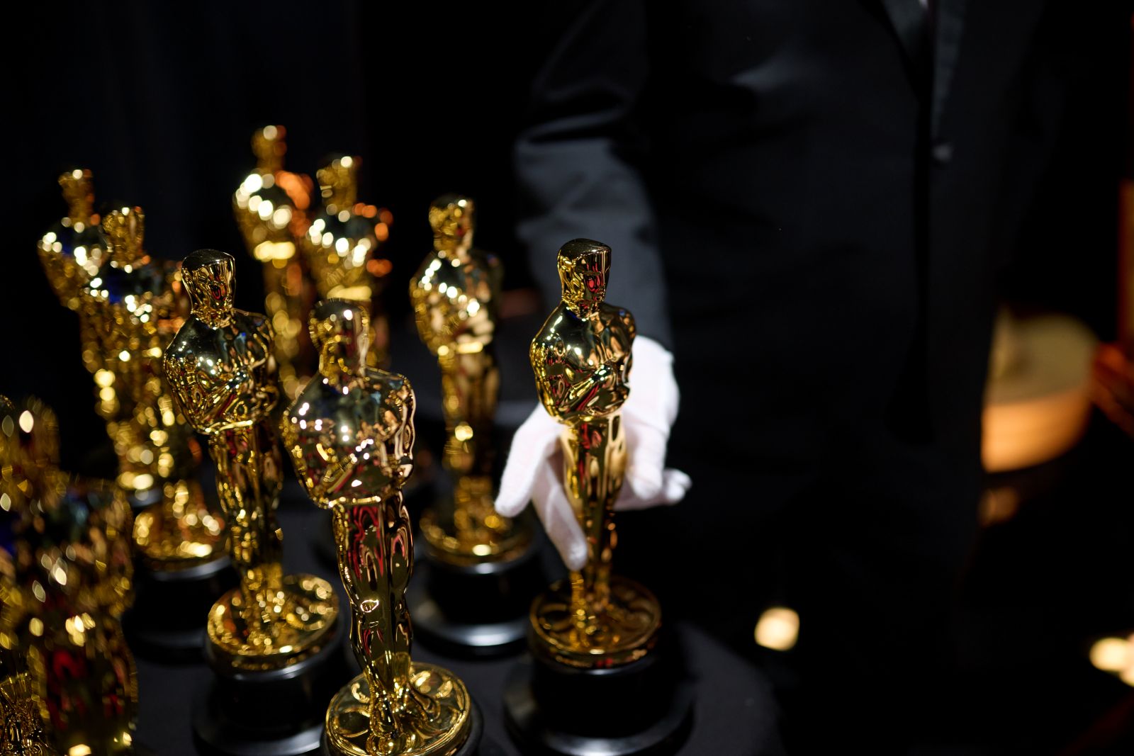 Two rows of Oscar statuettes stand backstage at the 2025 Academy Awards. A pair of hands in white gloves holds one statuette.
