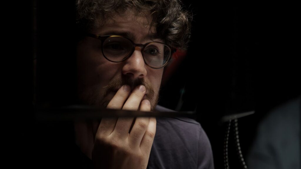 Filmmaker Daniel Roher is pictures in close-up. His hand is touching his face and he looks pensive while listening to an interviewee.