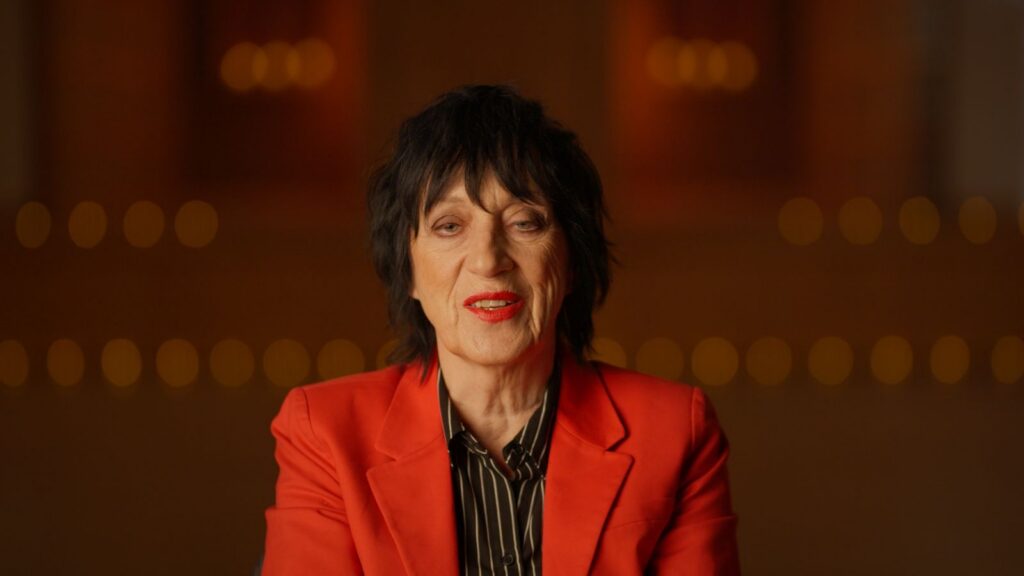 Rocker Carole Pope is seated for an interview. She is facing the camera in close-up and wearing a red blazer and black shirt.