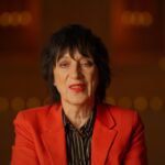 Rocker Carole Pope is seated for an interview. She is facing the camera in close-up and wearing a red blazer and black shirt.