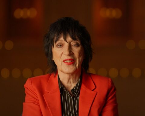 Rocker Carole Pope is seated for an interview. She is facing the camera in close-up and wearing a red blazer and black shirt.