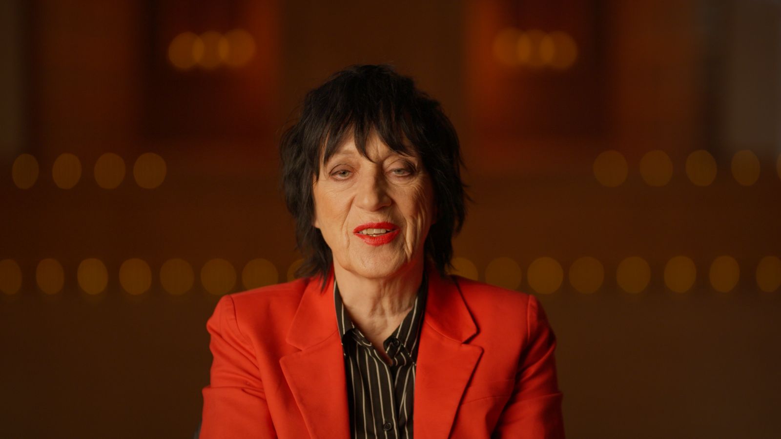 Rocker Carole Pope is seated for an interview. She is facing the camera in close-up and wearing a red blazer and black shirt.