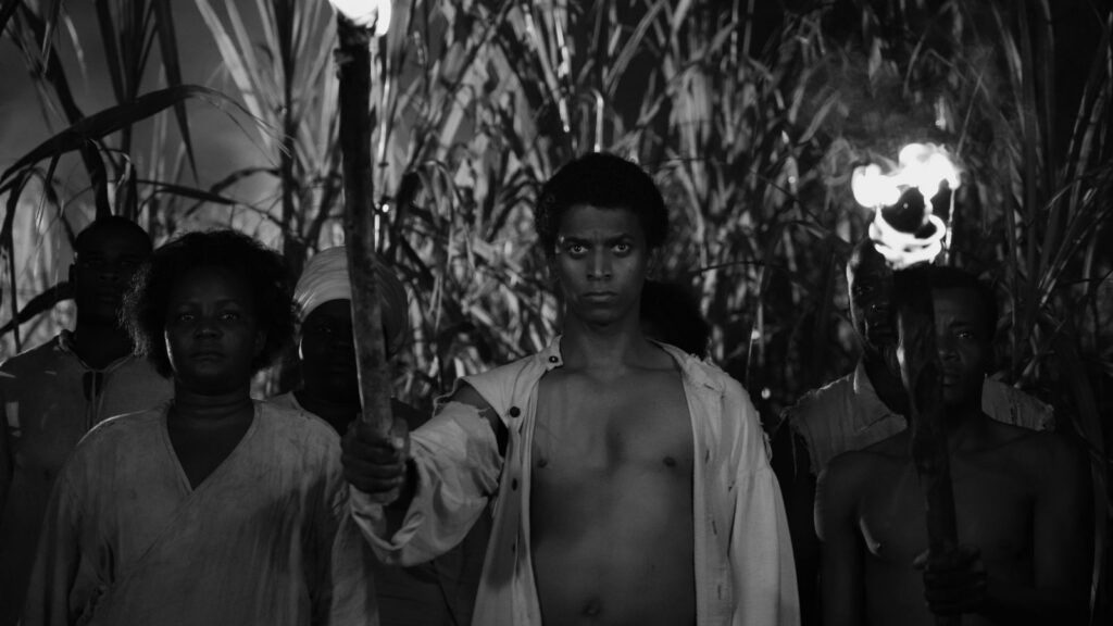 A black and white image of a Black Haitian man standing at the front of a line of people. He is wearing a white unbuttoned shirt and holding a lit torch.