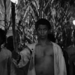 A black and white image of a Black Haitian man standing at the front of a line of people. He is wearing a white unbuttoned shirt and holding a lit torch.