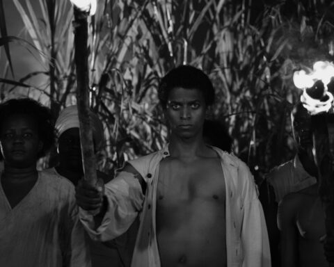 A black and white image of a Black Haitian man standing at the front of a line of people. He is wearing a white unbuttoned shirt and holding a lit torch.
