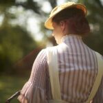 A woman stands with her back to the camera. She is wearing a red and white striped shirt and a straw hat. The sun beats down on her.