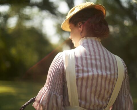 A woman stands with her back to the camera. She is wearing a red and white striped shirt and a straw hat. The sun beats down on her.