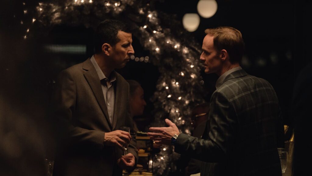 Two men are having an argument. They are both wearing dark suits and are standing in front of a Christmas tree with white lights.