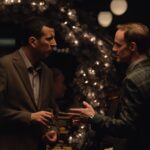 Two men are having an argument. They are both wearing dark suits and are standing in front of a Christmas tree with white lights.