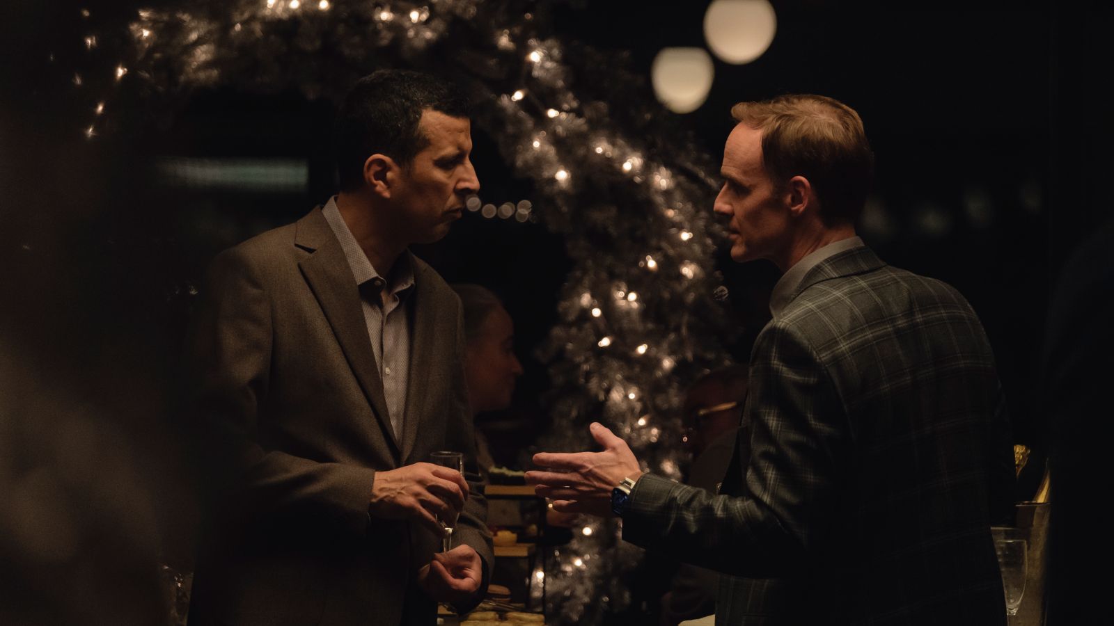 Two men are having an argument. They are both wearing dark suits and are standing in front of a Christmas tree with white lights.