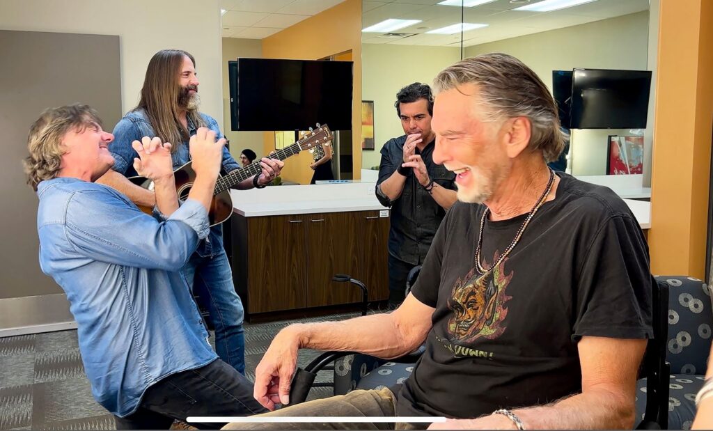 Musician Kenny Loggins sits in a chair in a dressing room. He is wering a black t-shirt and laughing. Three friends are in the background, also laughing.