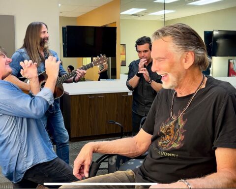 Musician Kenny Loggins sits in a chair in a dressing room. He is wering a black t-shirt and laughing. Three friends are in the background, also laughing.