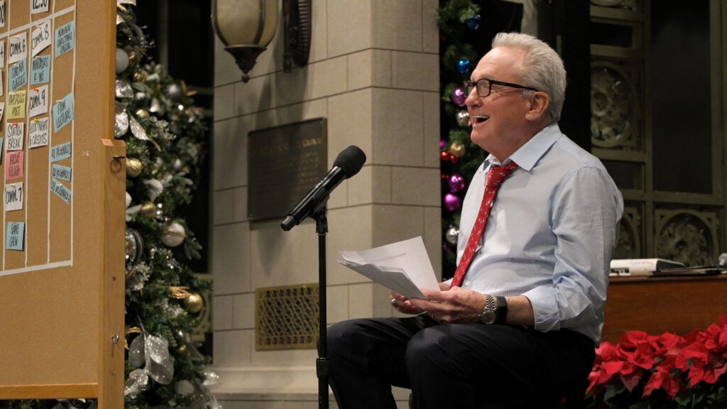 Producer Lorne Michaels sits at a microphone delivering a speech and laughing. He is wearing a white shirt and red tie.