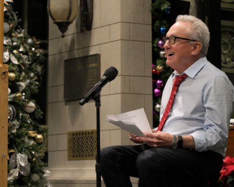 Producer Lorne Michaels sits at a microphone delivering a speech and laughing. He is wearing a white shirt and red tie.