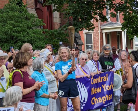 79-year-old runner Katrine Switzer holds the Olympic torch. She is surrounded by an enthusiastic crowd.