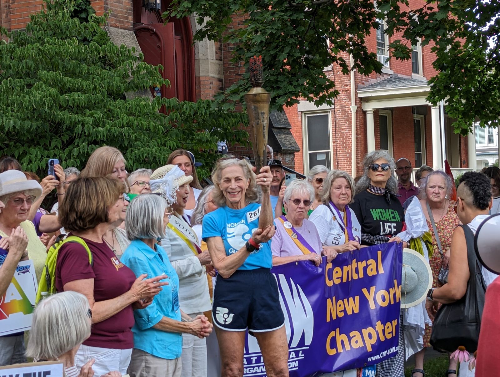 79-year-old runner Katrine Switzer holds the Olympic torch. She is surrounded by an enthusiastic crowd.