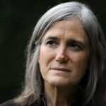 Journalist Amy Goodman is pictured in close-up. She is a white woman in her 60s with grey hair and brown eyes. She is pictured against a black backdrop and her eyes are looking at something to screen left.