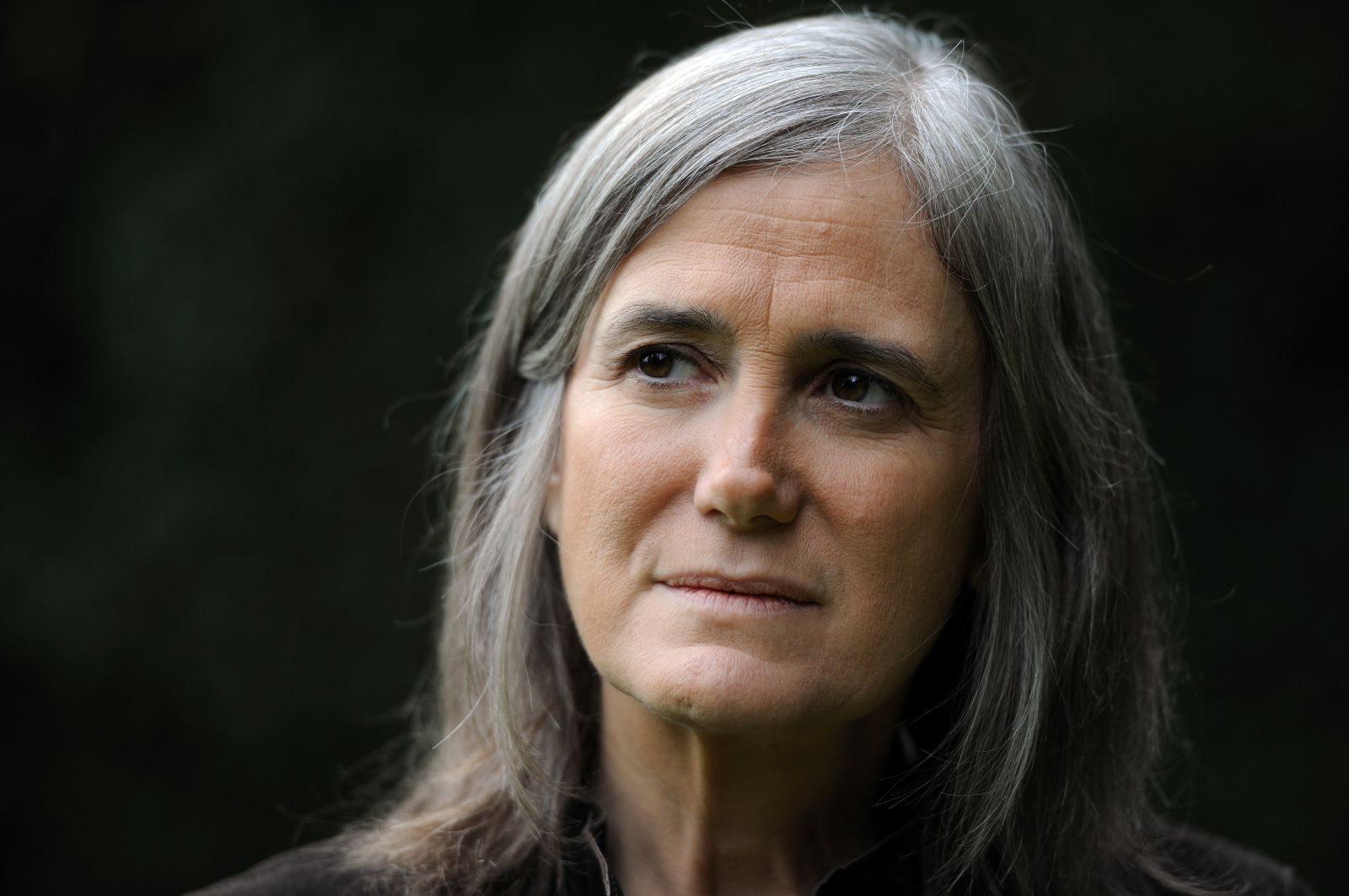 Journalist Amy Goodman is pictured in close-up. She is a white woman in her 60s with grey hair and brown eyes. She is pictured against a black backdrop and her eyes are looking at something to screen left.