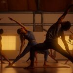 A group of people perform yoga stretches. The are pictured in a golden hue.