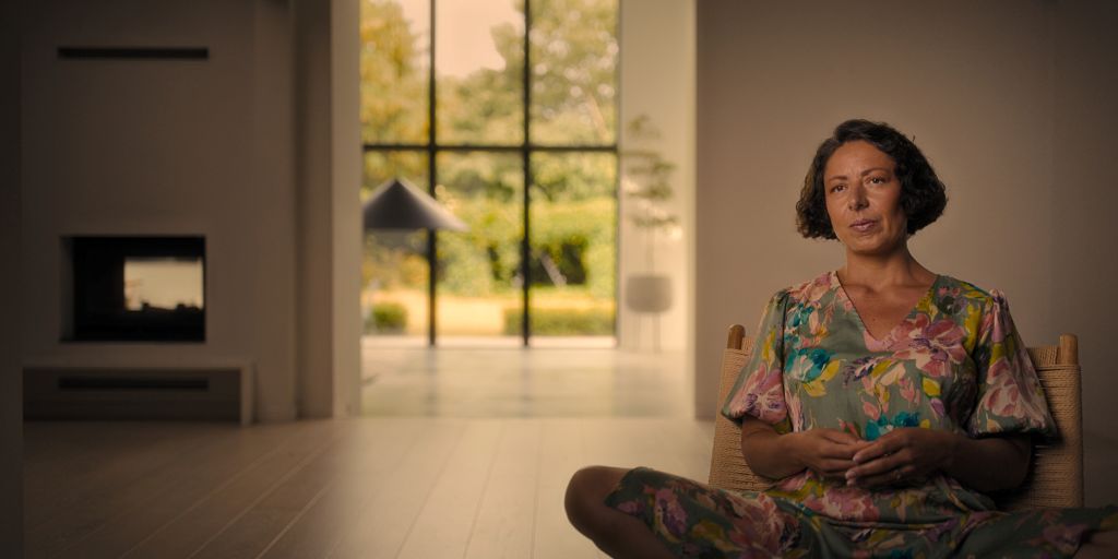 A woman sits cross-legged in a chair. She is wearing a floral print shirt and black leggins. She has short brown hair and is facing the camera. The room has a lot of natural light.